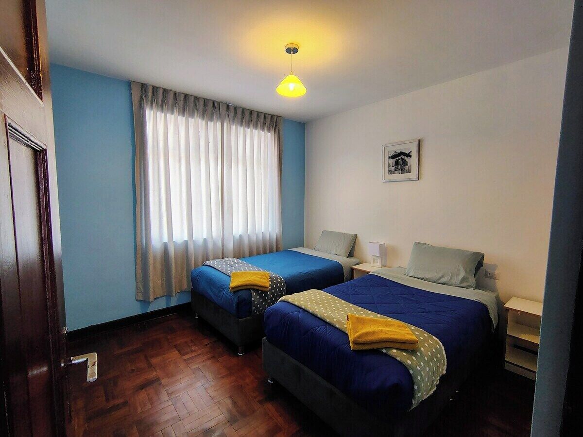 A room with two beds, a window with curtains, a wall-mounted picture, and a wooden floor.