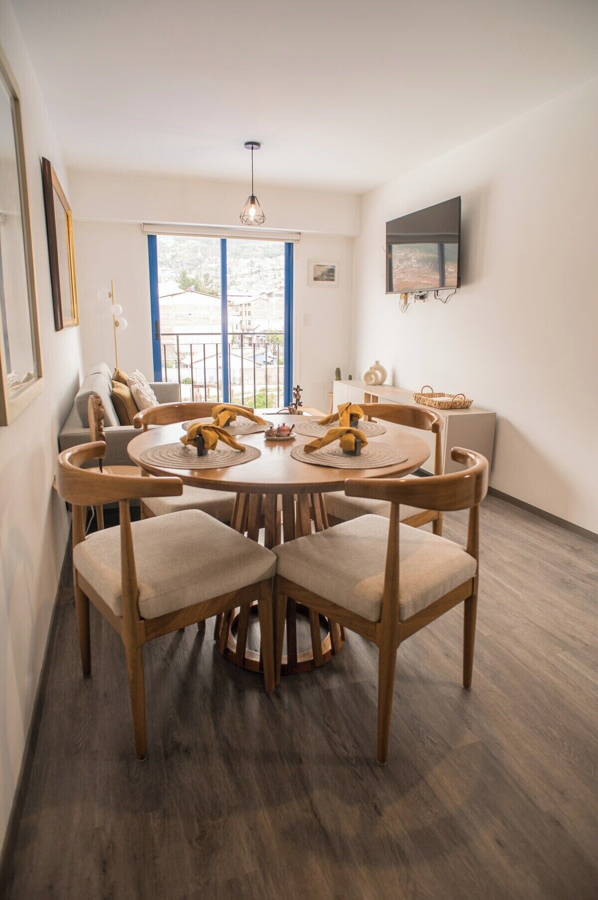 A dining area with a round table and chairs, a TV mounted on the wall, and a view through the window.