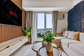 Een moderne woonkamer met een grote flatscreen-tv, een bank, een salontafel en een stadsgezicht door een groot raam.