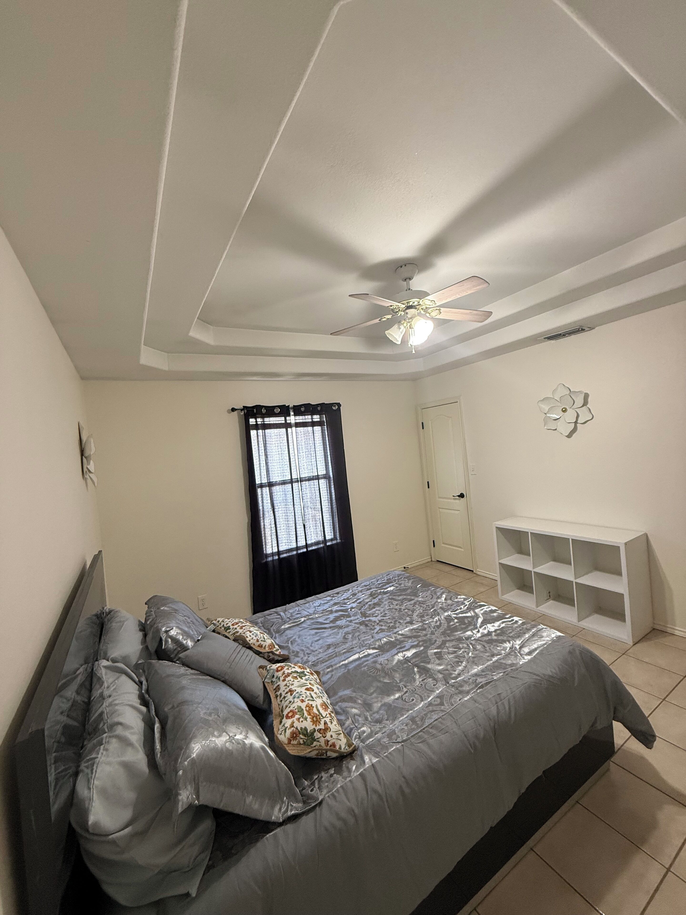 Un dormitorio con cama, ventilador de techo, ventana con cortinas, armario blanco y estante en la pared.