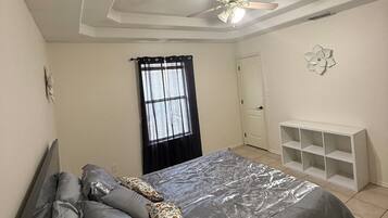Un dormitorio con cama, ventilador de techo, ventana con cortinas, armario blanco y estante en la pared.