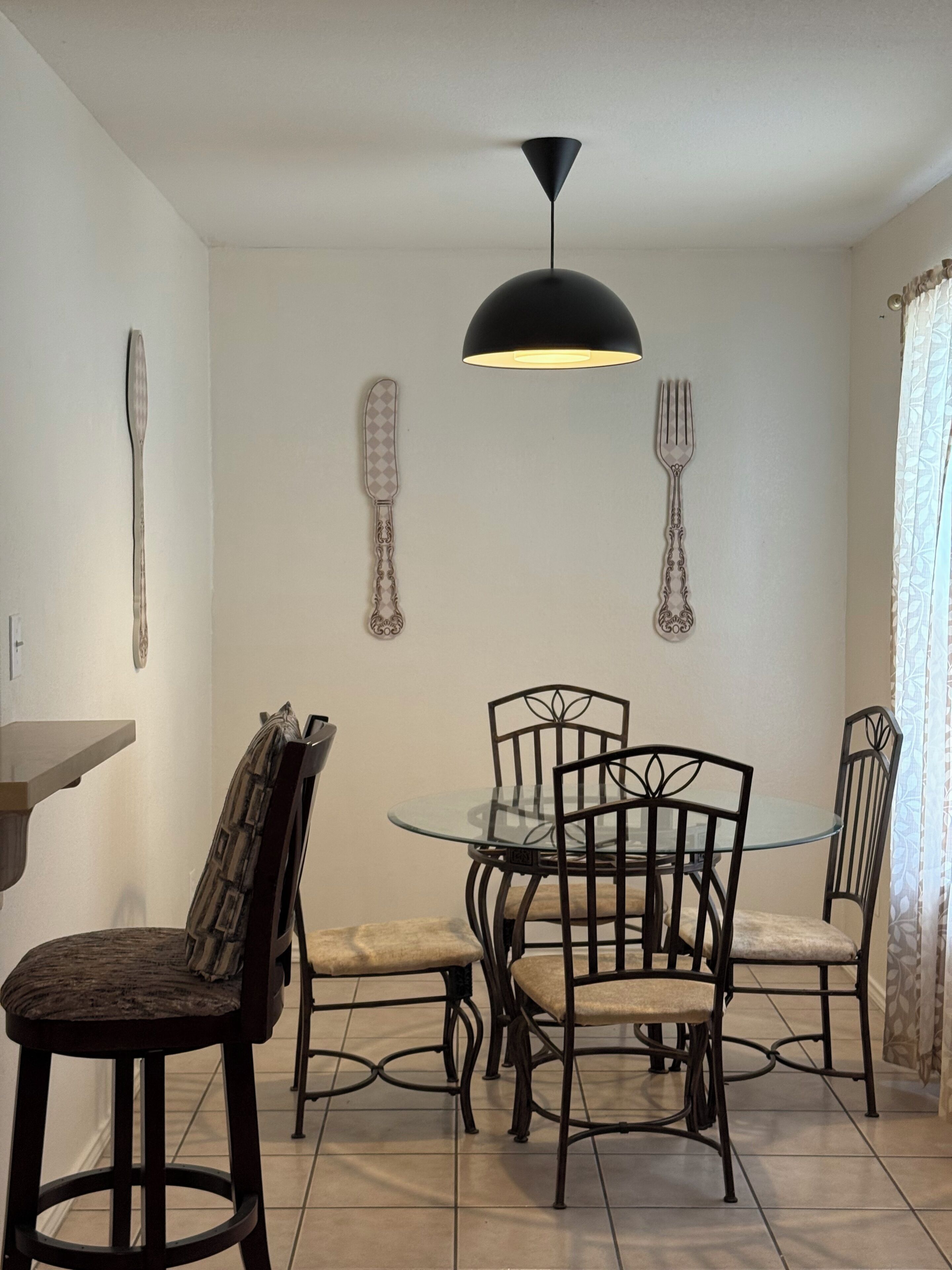 A dining area with a round glass table, four chairs, a black pendant light, and wall-mounted cutlery.