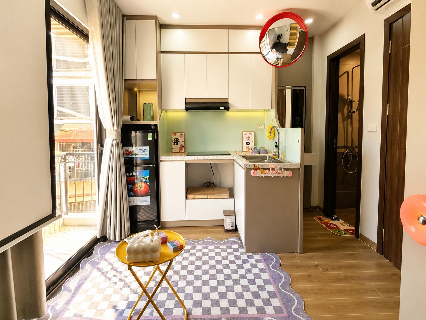 A compact kitchen with a refrigerator, microwave, and sink. There's a small dining area with a checkered rug and a yellow table.