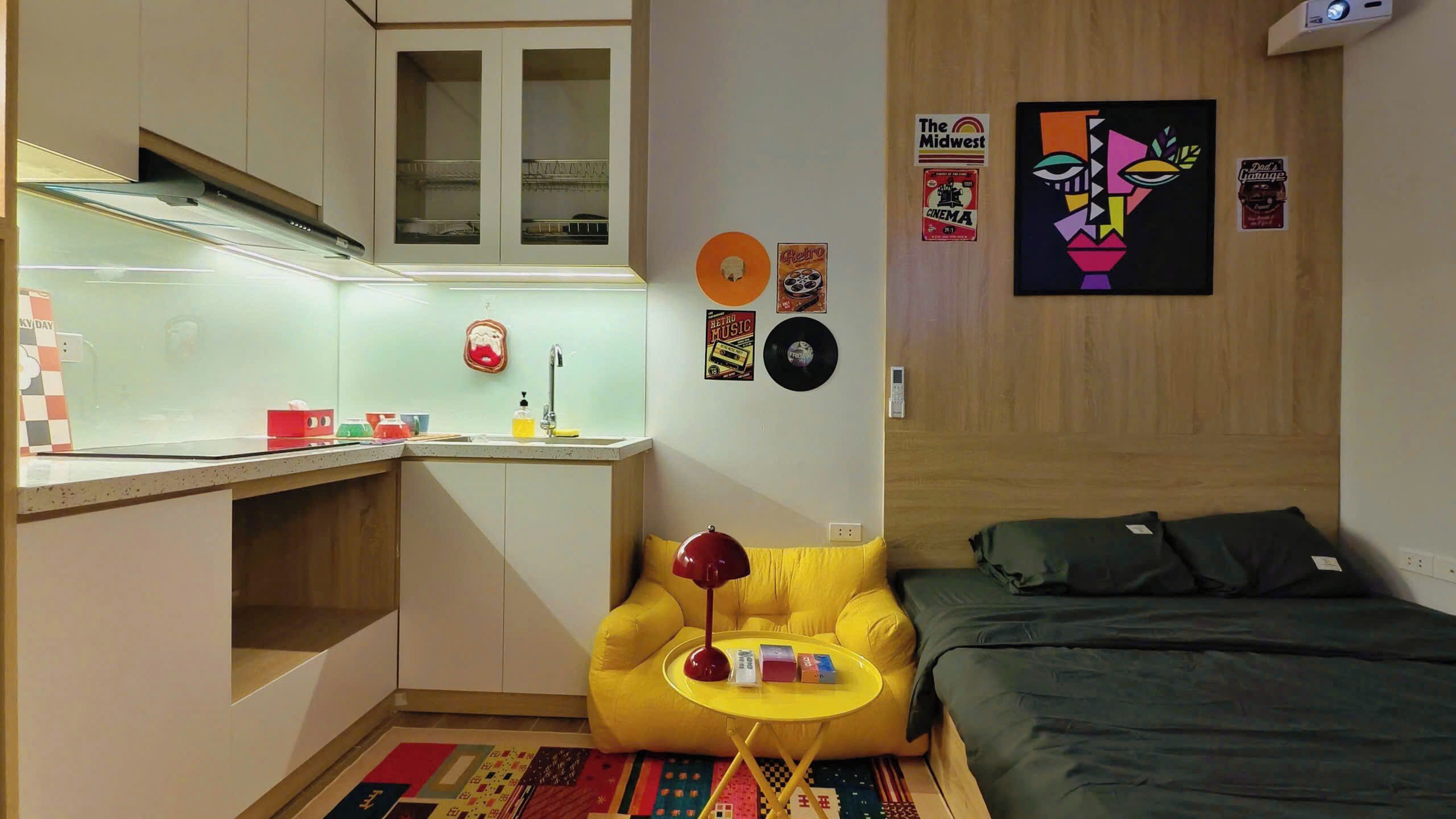 A compact room with a kitchenette, a bed, a yellow armchair, and various wall decorations.