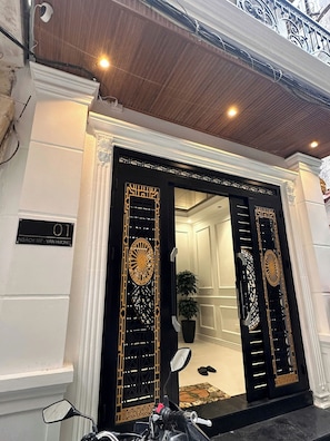 Een hotelentree met een decoratieve deur in zwart en goud, een bord met kamer nummer 01 en een potplant binnen.