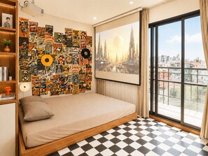 Un dormitorio con piso a cuadros, una cama con un almohadón, una estantería, una pared con varios carteles y un ventanal con vista a la ciudad.