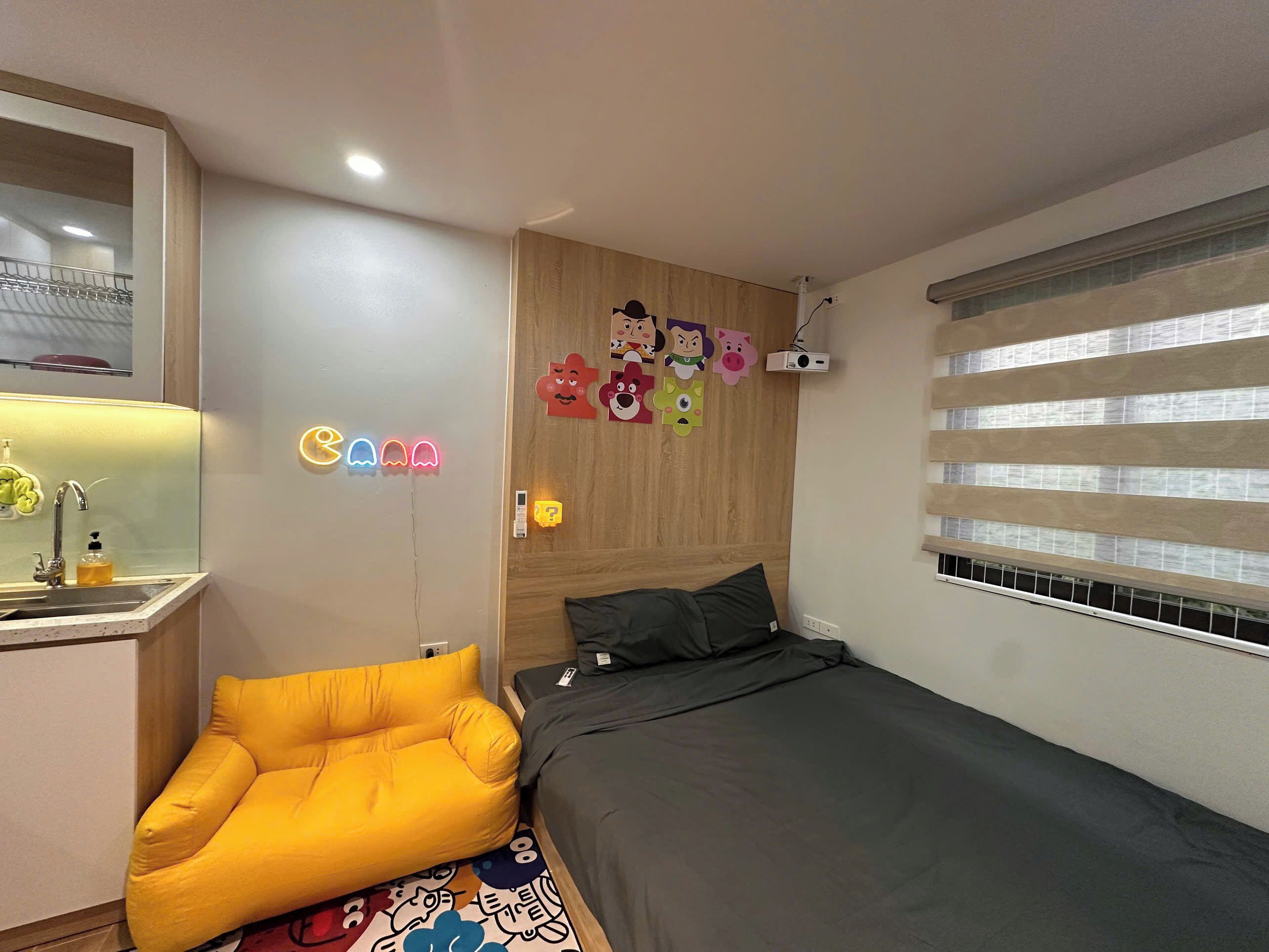 A bedroom with a bed, a yellow chair, a bedside table, a mirror, and a window with blinds.