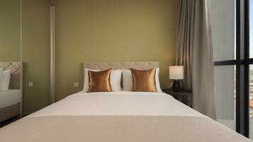 Een hotelkamer met een groot bed, hoofdeinde, twee kussens, een nachtkastje met lamp en uitzicht door een raam.