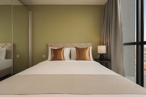 A hotel room with a large bed, a headboard, two pillows, a nightstand with a lamp, and a view through a window.