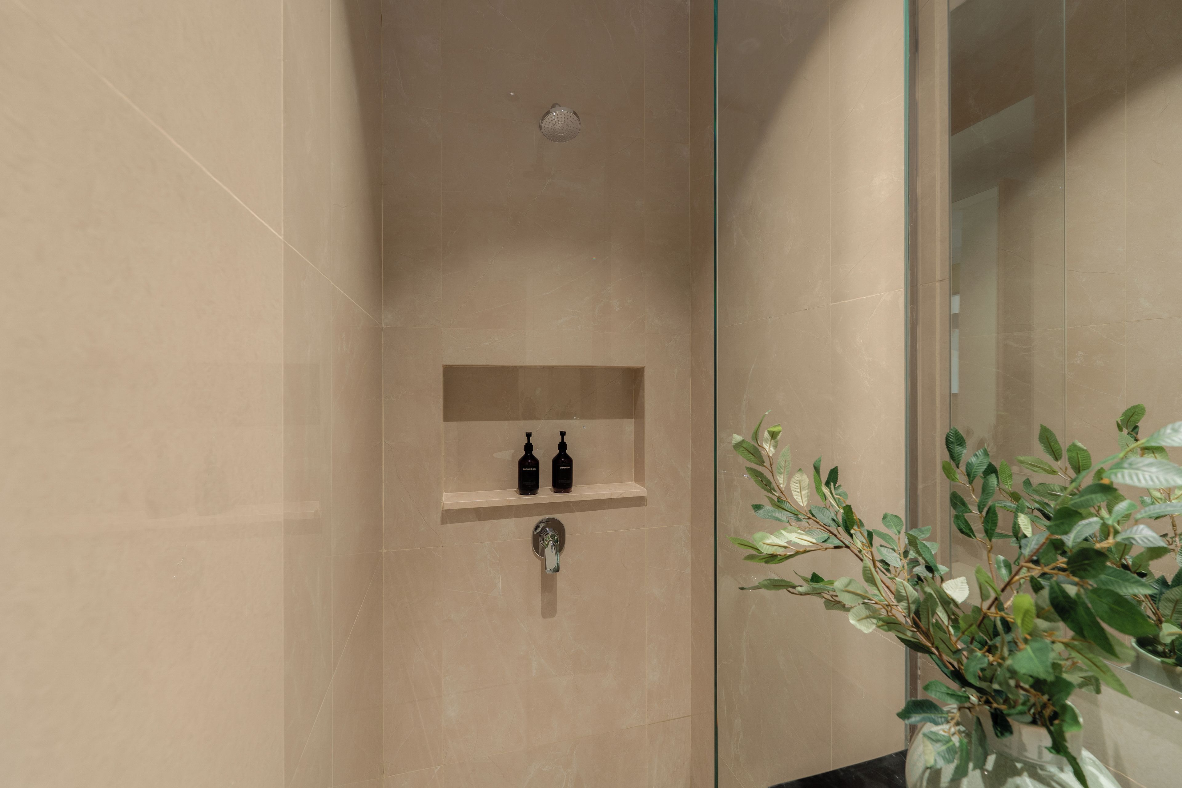Un baño con mampara de ducha de vidrio, un estante empotrado con dos botellas y una planta en maceta.