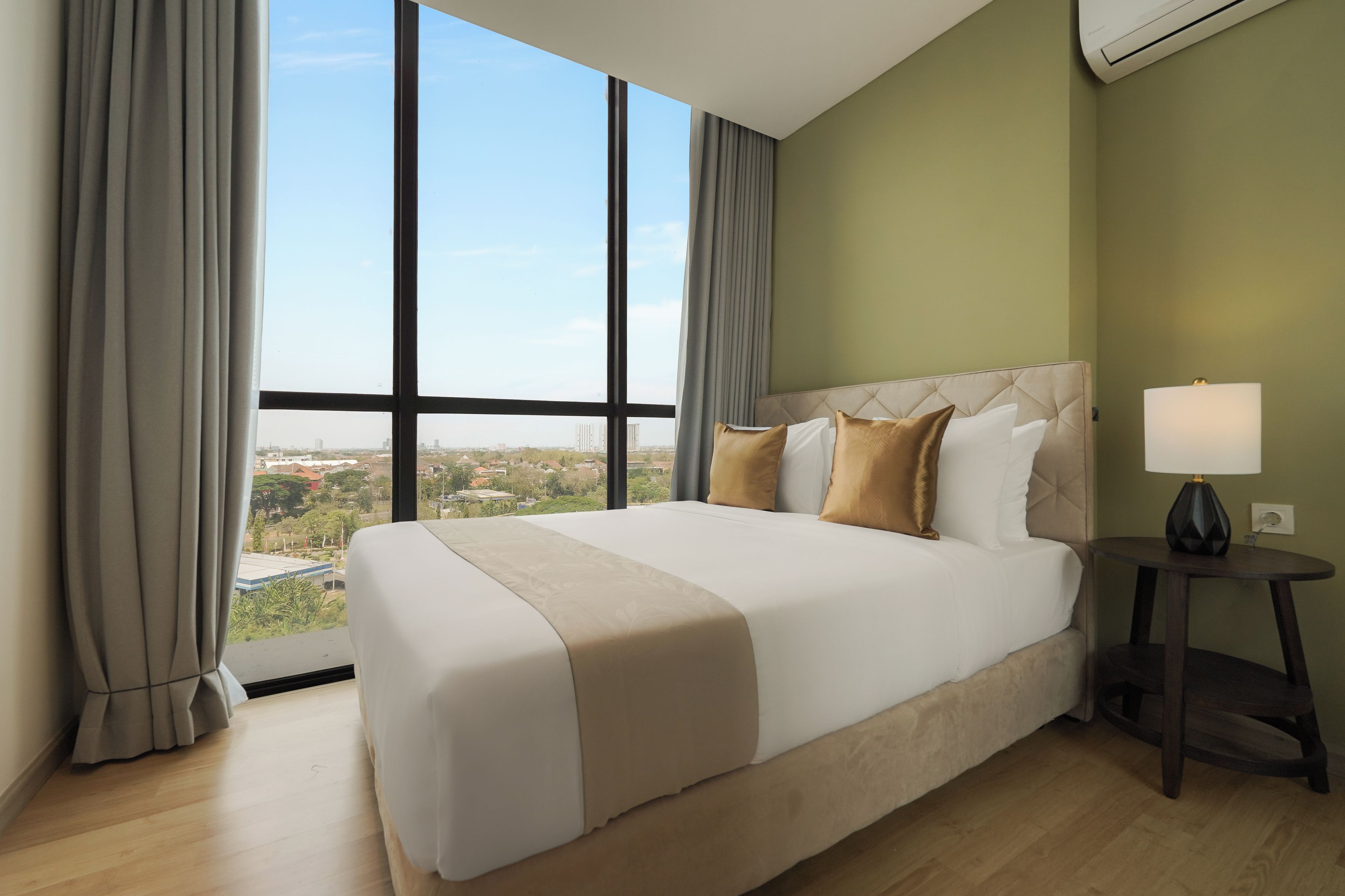 A hotel room with a large bed, a nightstand, a lamp, and a view of a cityscape through large windows.