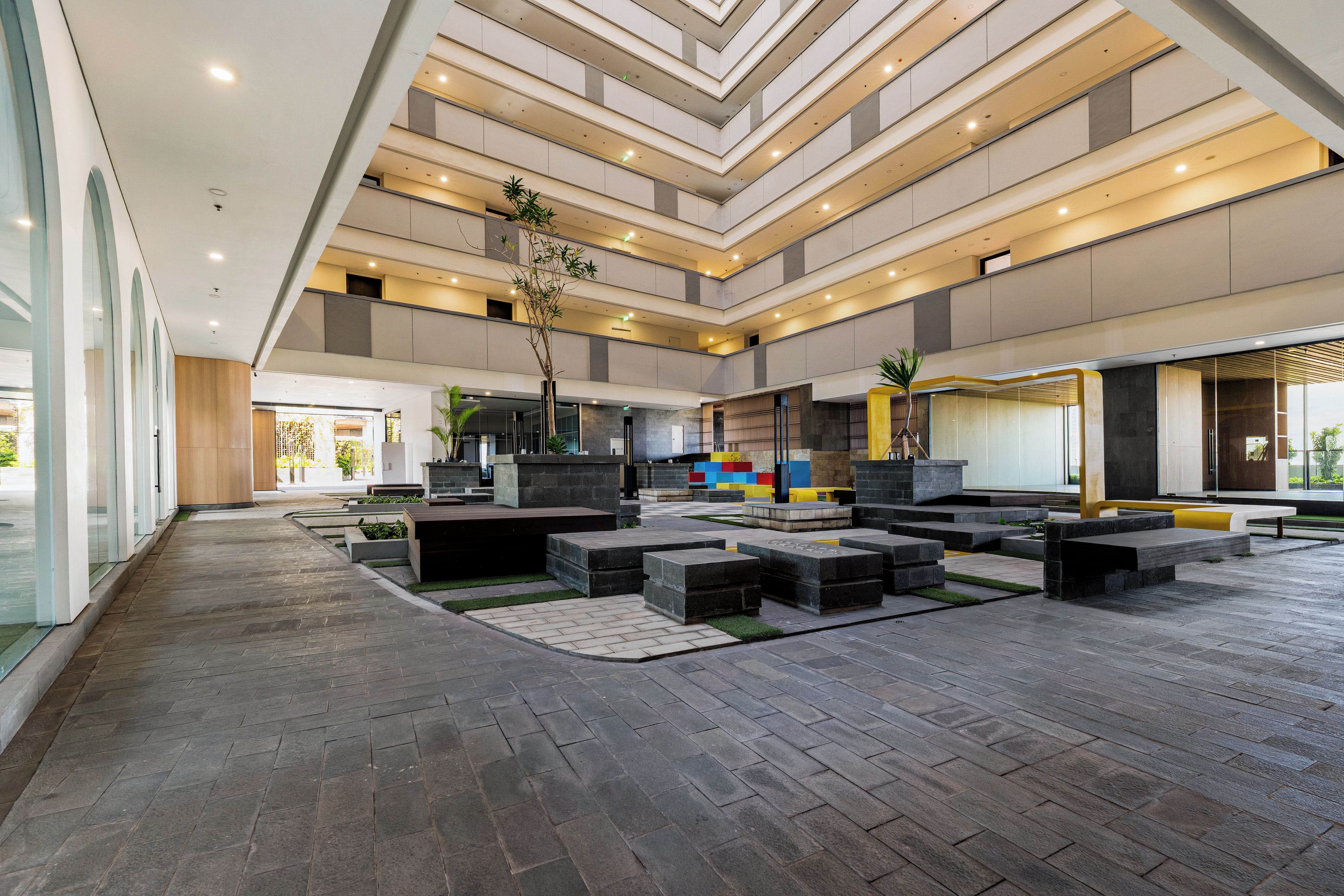 A spacious atrium with a modern design, featuring a central open area with seating and planters, and a multi-level ceiling with recessed lighting.