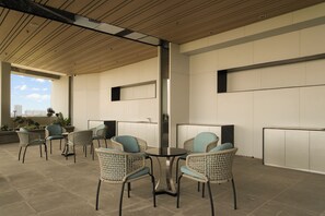 Uma área de estar moderna ao ar livre, com cadeiras de vime e uma mesa redonda, com vista para a cidade através de uma grande janela.