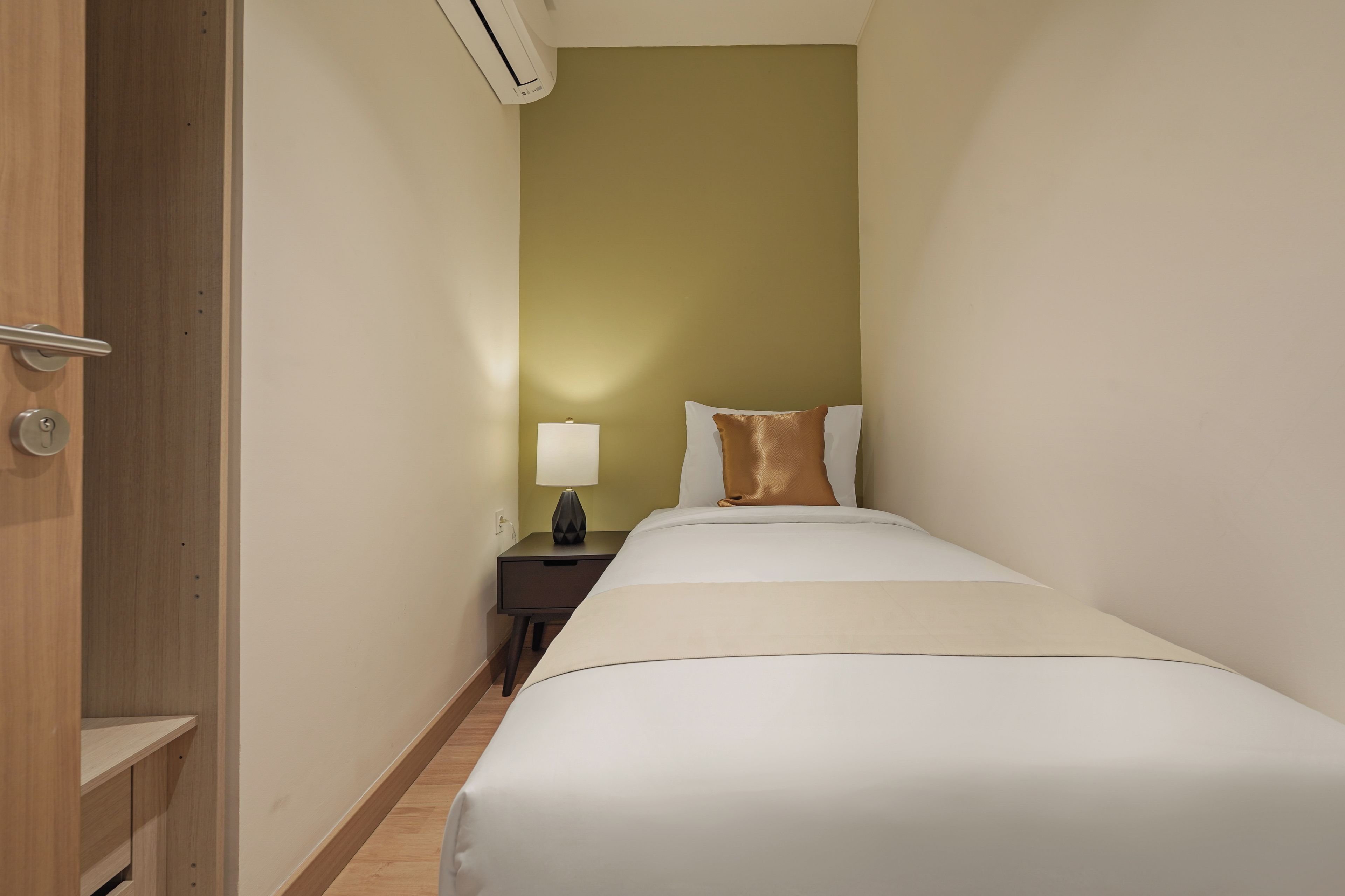 A narrow hotel room with a single bed, a nightstand, a lamp, and a door with an air conditioning unit.