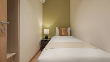 A narrow hotel room with a single bed, a nightstand, a lamp, and a door with an air conditioning unit.