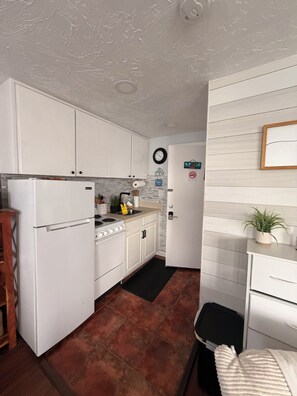Une cuisine de taille réduite avec des meubles blancs, une cuisinière et un réfrigérateur. Un tapis noir est posé sur le sol, et une plante se trouve sur le plan de travail.