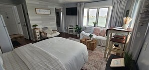 Ein Hotelzimmer mit einem großen Bett, einem Schreibtisch mit Computer, einem Sessel, einem Fenster mit Blick auf das Wasser und einem Schild an der Wand.
