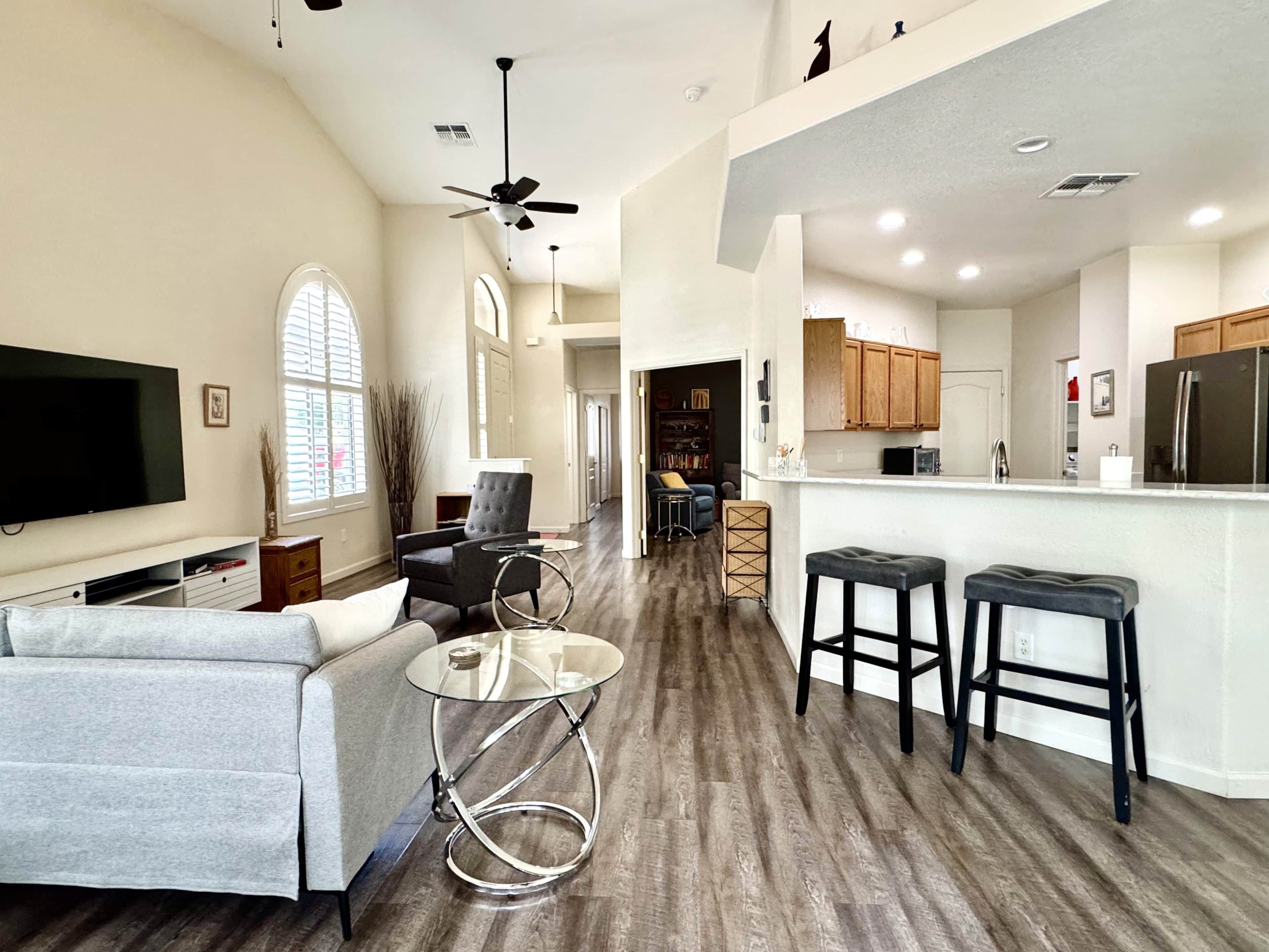 A spacious living area with a flat-screen TV, a grey sofa, a glass coffee table, and a ceiling fan.