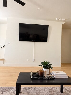Un salon moderne avec un téléviseur à écran plat fixé au mur, une table basse agrémentée d’une plante et de livres, et un plancher en bois.