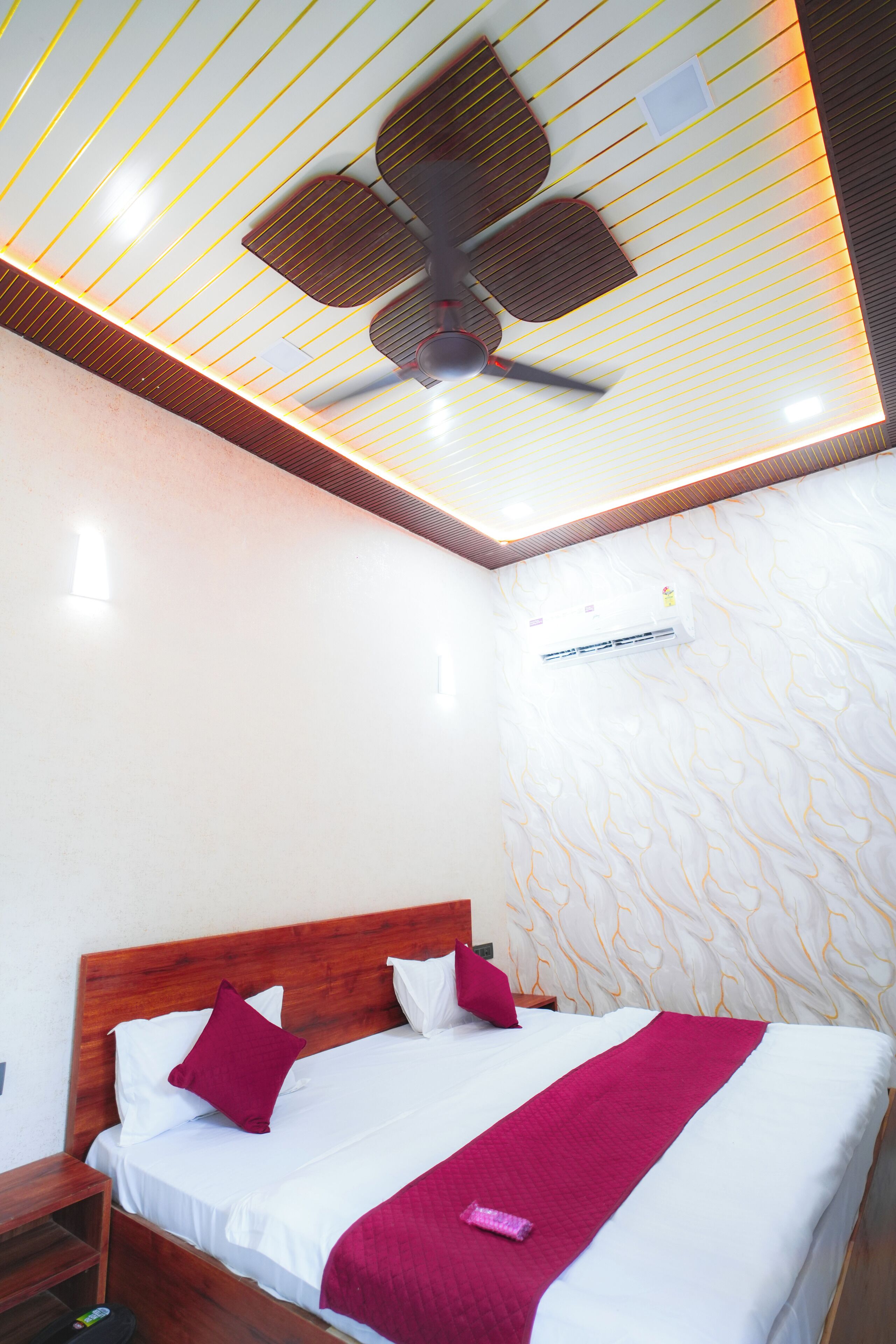 A hotel room with a bed, wooden headboard, white bedding, a ceiling fan, and a patterned wall.