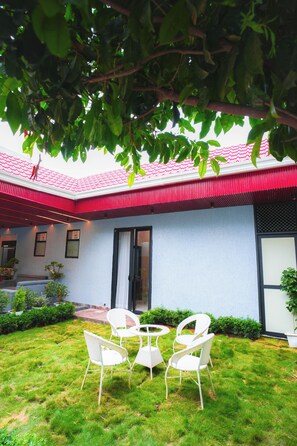 A patio with white chairs and a table, a red roof, and a white wall with a door. - Panther Farm Patrakar (Jaipur)