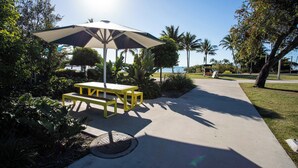 A sunny outdoor area with a picnic table and umbrella, surrounded by greenery and palm trees. - Marina Views at Airlie 10H (Airlie Beach)