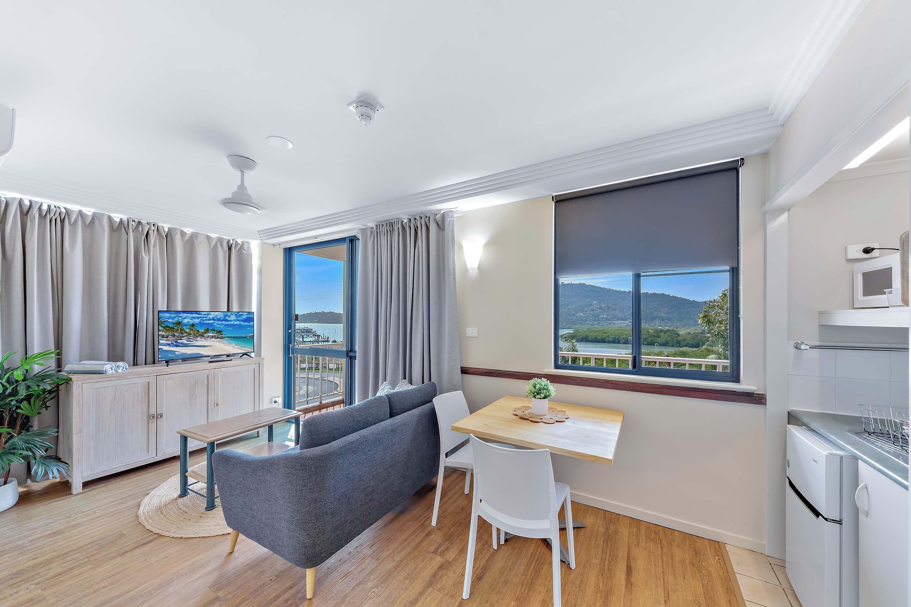 A modern living room with a sofa, a dining table, and a TV. There are large windows offering a view of the outdoors.