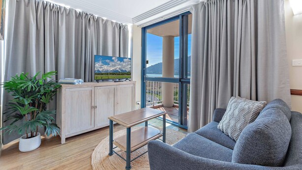 A modern living room with a sofa, a coffee table, a TV on a cabinet, and a view of mountains through the glass door. - Marina Views at Airlie 10H (Airlie Beach)