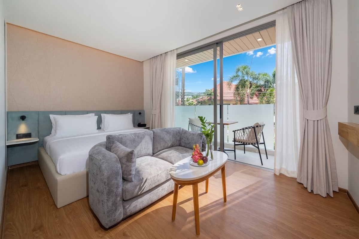 Habitación de hotel con una cama, un sofá gris, una mesita con frutas y vistas a través de un ventanal corredero.