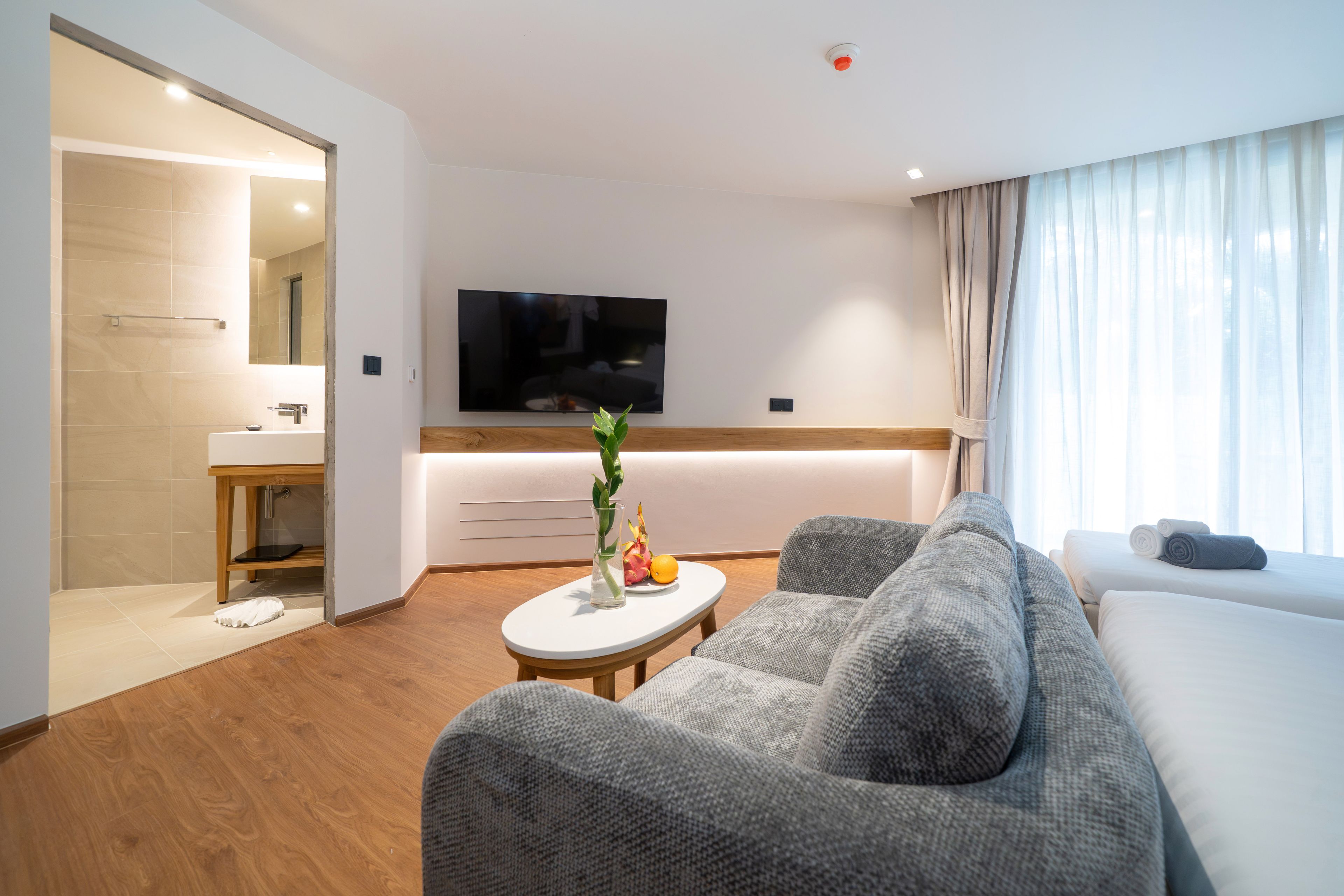 A modern hotel room with a sofa, a flat-screen TV, a small table with a vase and fruit, and a bathroom visible through an open door.