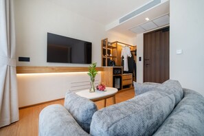 A modern living room with a large gray sofa, a small round table with a vase and a plant, a flat-screen TV mounted on the wall, and a wooden cabinet with a robe hanging. - The Beachside By Himmapana (Kamala)