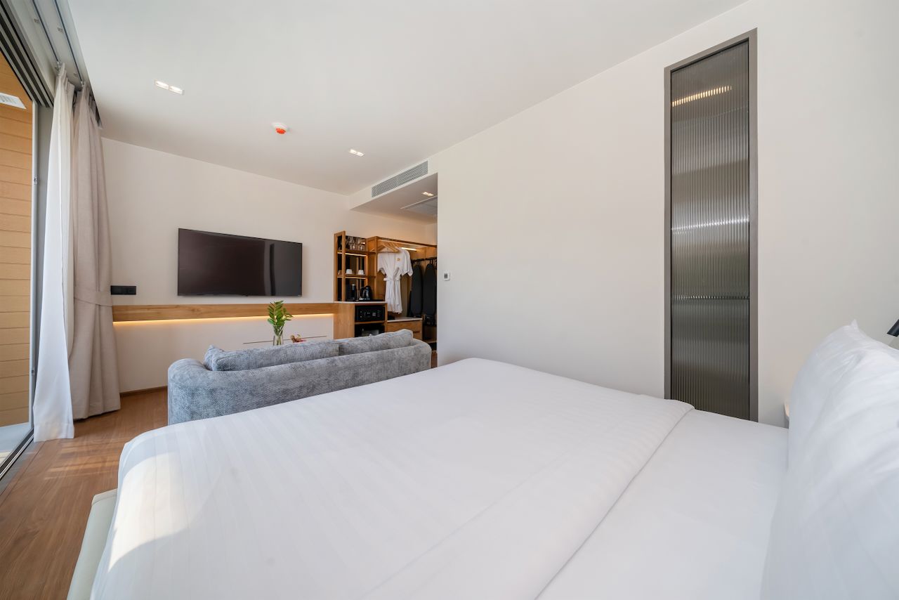 A modern hotel room with a large bed, a flat-screen TV mounted on the wall, a seating area with a sofa, and a wooden wardrobe.
