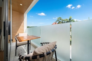 Un balcón con una mesa y sillas de madera, una barandilla de vidrio y vistas al cielo y edificios a la distancia.