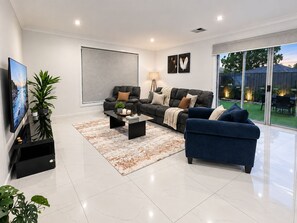 Ein modernes Wohnzimmer mit Flachbildfernseher, zwei Sofas, einem Couchtisch und einem großen Fenster mit Blick auf einen Garten.