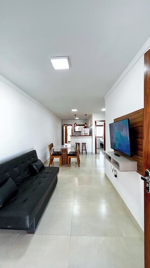 A modern living room with a black sofa, a dining area with a table and chairs, a kitchen in the background, and a TV mounted on a wooden wall. - Complete house 900m from Geribá Beach, with balcony and barbecue in Búzios. (Armação dos Búzios)