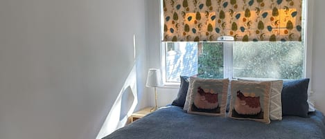 A bedroom with a large bed, a window with a patterned blind, and a small bedside table with a lamp.