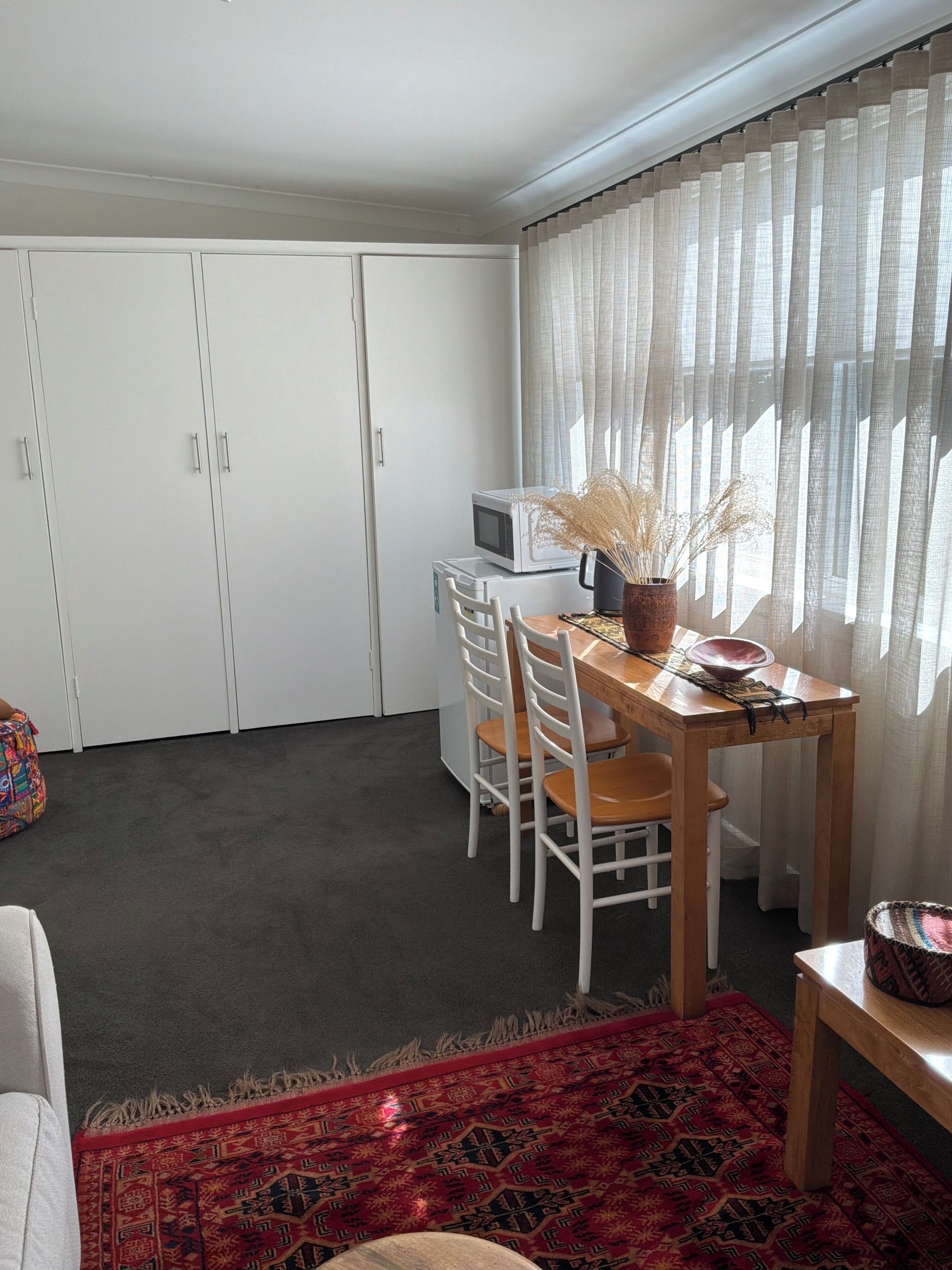A room with a dining table, chairs, a microwave, a small rug, and a window with sheer curtains.