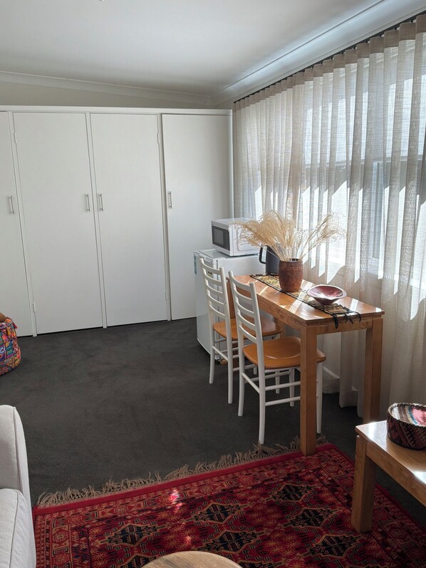 A room with a dining table, chairs, a microwave, a small rug, and a window with sheer curtains.