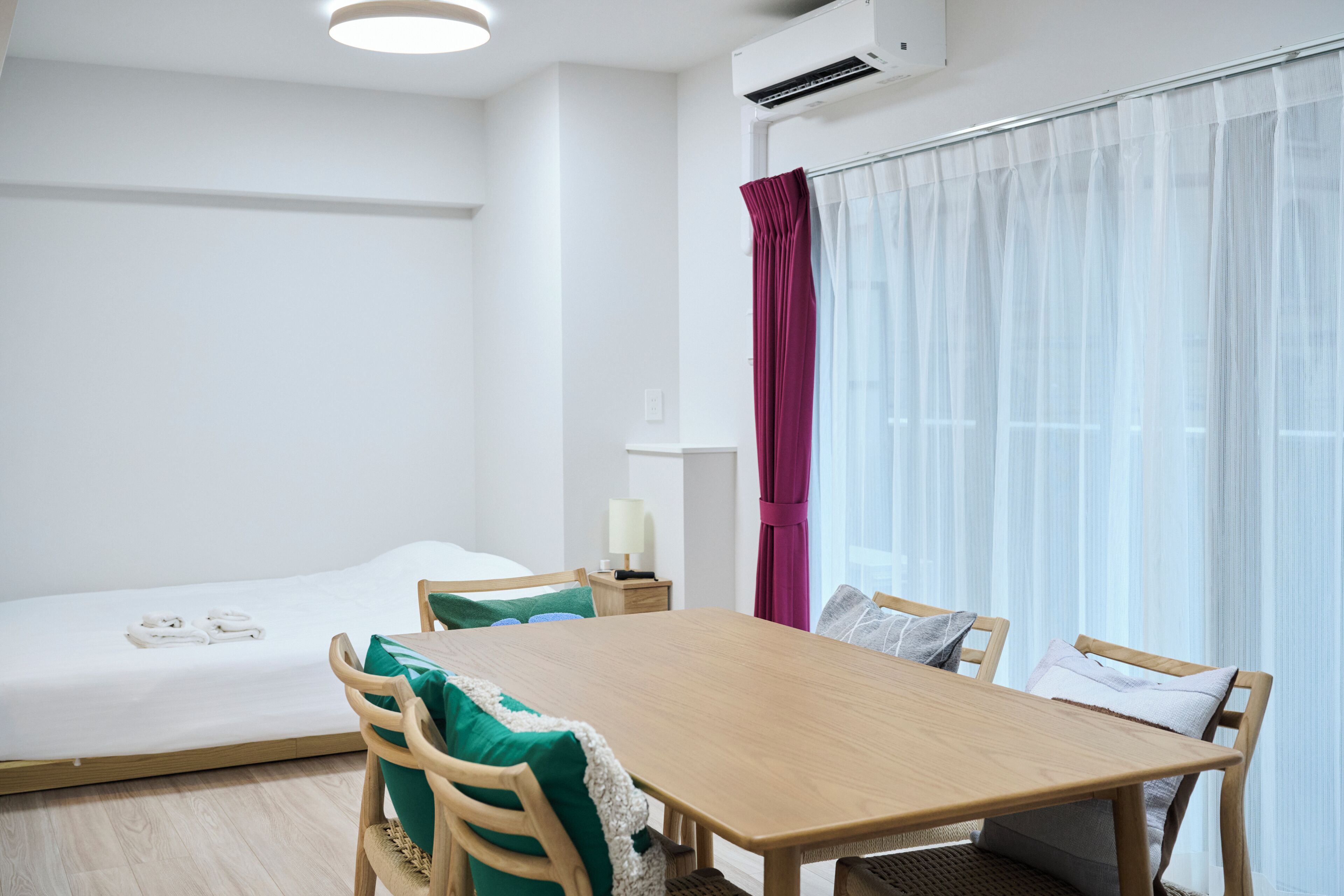 A hotel room with a bed, a wooden table, and chairs. There is a window with sheer curtains and a red curtain partially drawn.