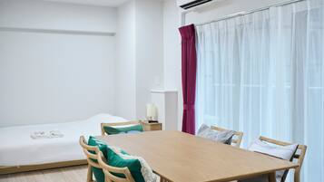 A hotel room with a bed, a wooden table, and chairs. There is a window with sheer curtains and a red curtain partially drawn.