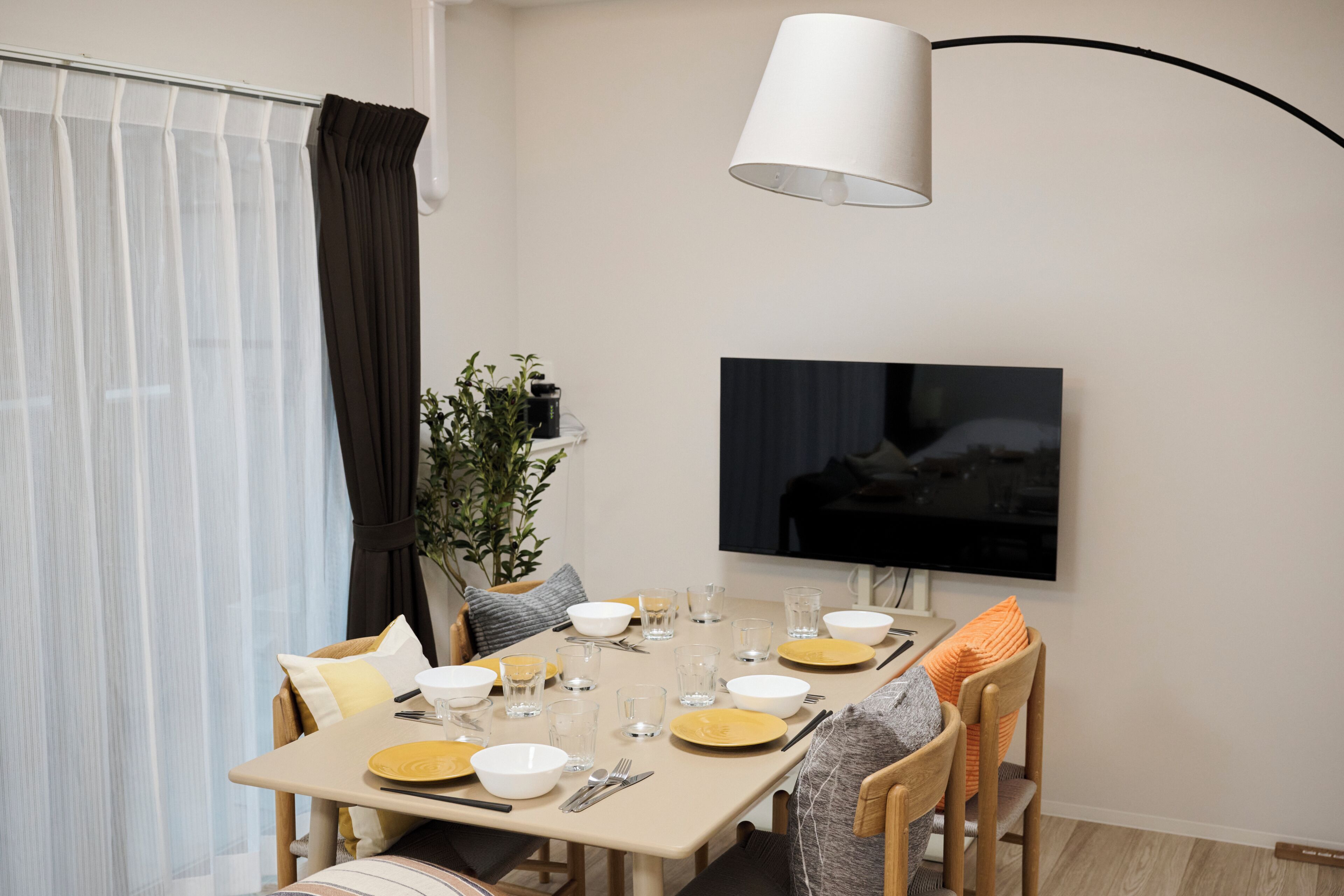 Uma sala de jantar com uma mesa posta para dois, uma televisão fixada na parede e uma planta sobre a mesa.