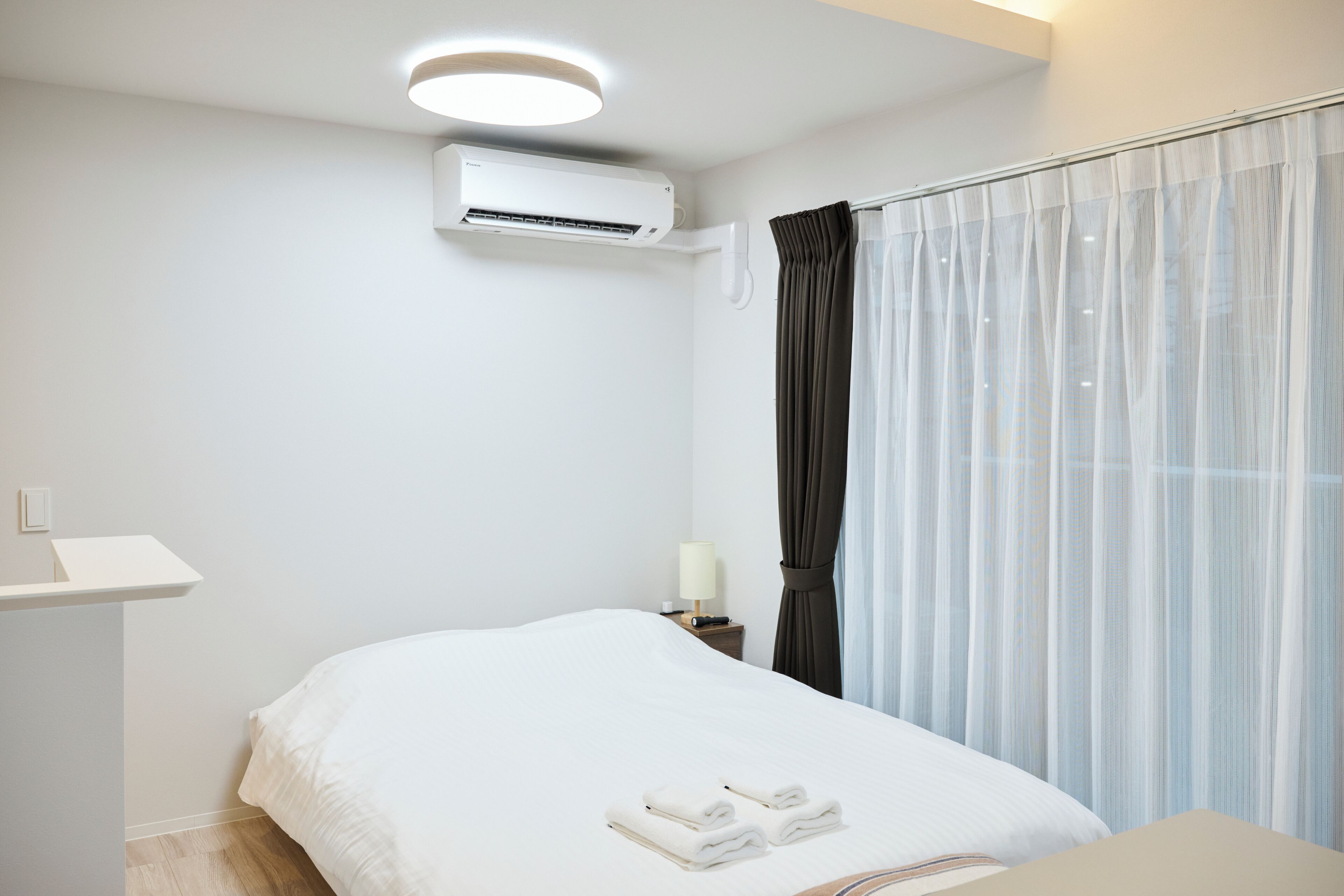 A hotel room with a bed, an air conditioning unit, a bedside table with a lamp, and sheer curtains.