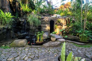 Un sendero de piedra lleva a un pequeño estanque con cascada, rodeado de exuberante vegetación y una estructura rústica de piedra con un arco.