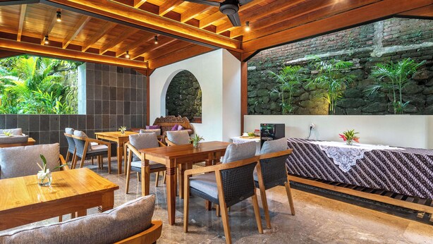 A restaurant with wooden tables and chairs, a stone wall with greenery, and a ceiling fan.