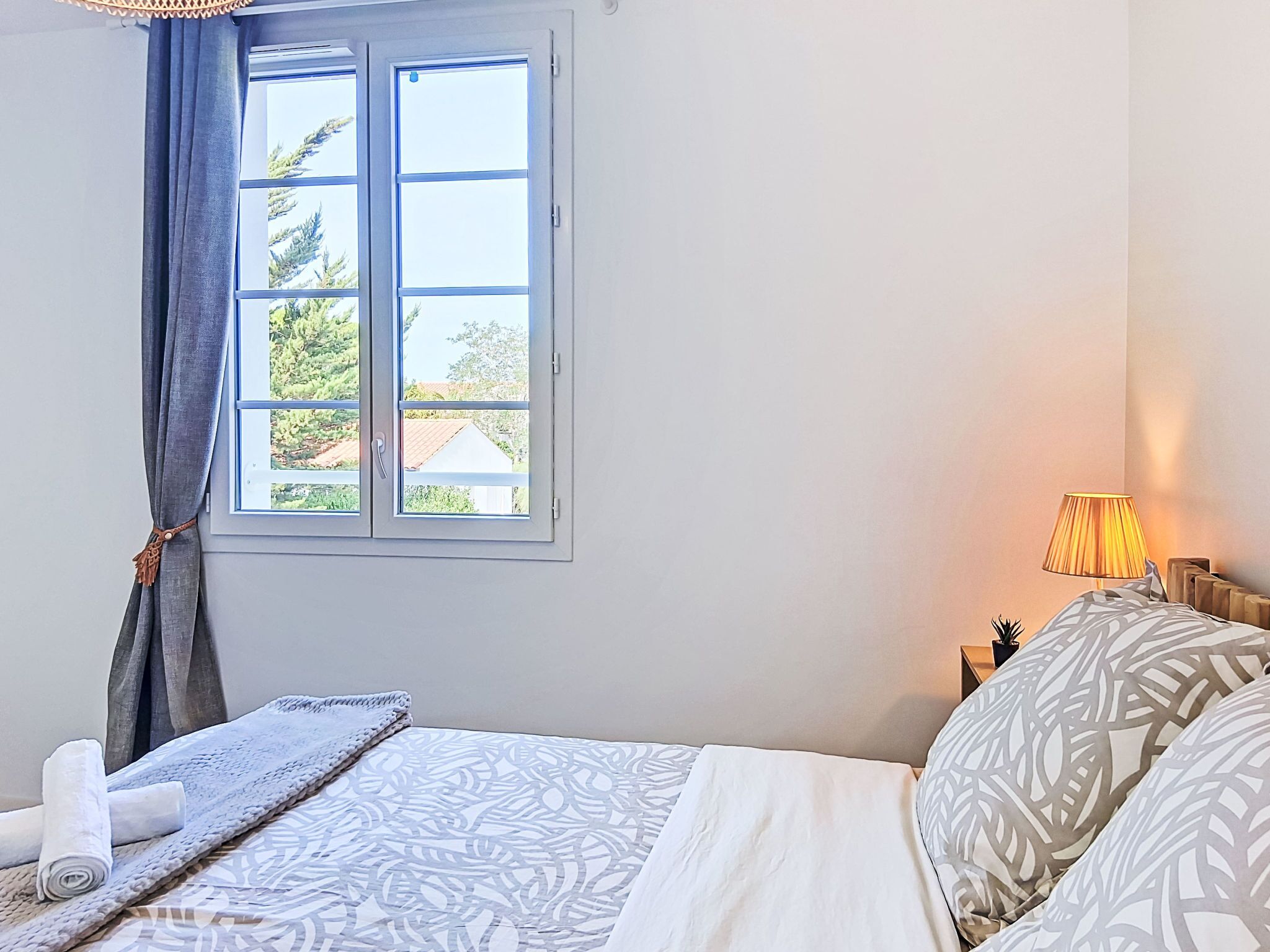 Een slaapkamer met een bed, een raam, een lamp en uitzicht op bomen en gebouwen.