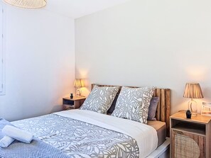 Uma cama bem arrumada com roupa de cama estampada, uma mesa de cabeceira com abajur e uma pequena planta em vaso.