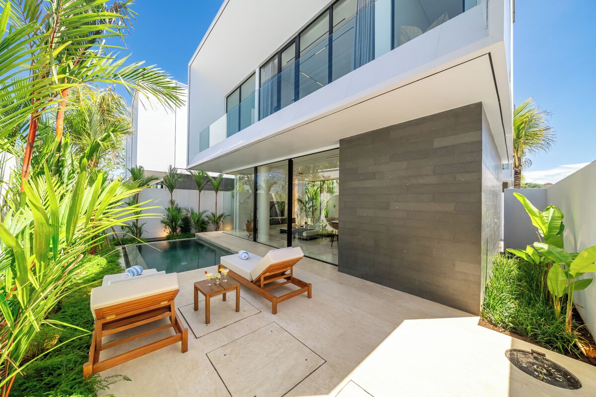 A modern outdoor pool area with lounge chairs and a table, surrounded by tropical plants.