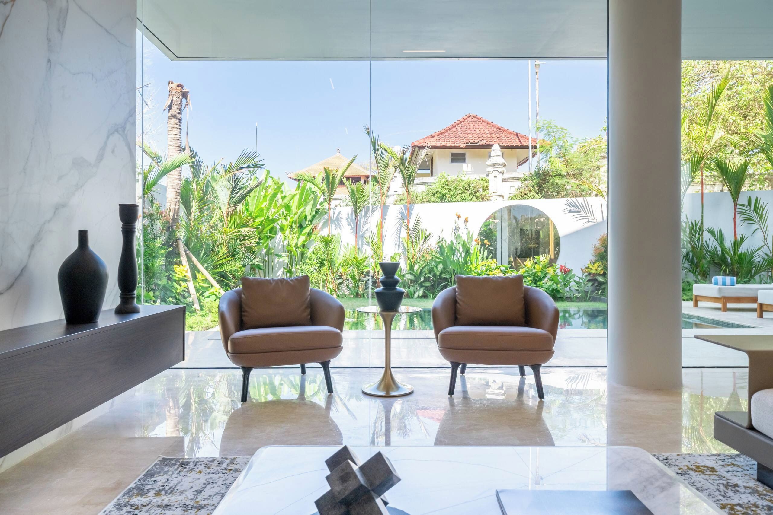 Una sala de estar moderna con pared de vidrio, dos sofás de cuero, una mesa de centro y vistas a una piscina y follaje.