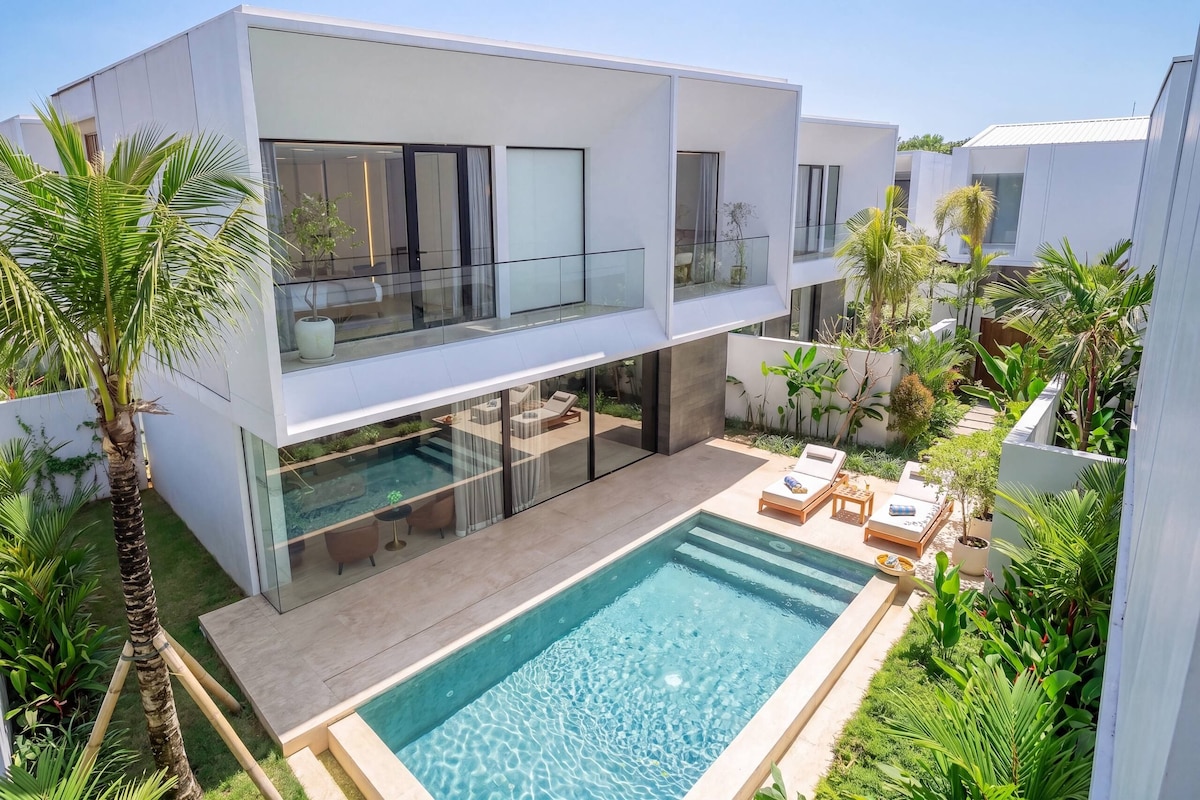 A modern two-story house with a glass balcony, a swimming pool, and lush greenery.