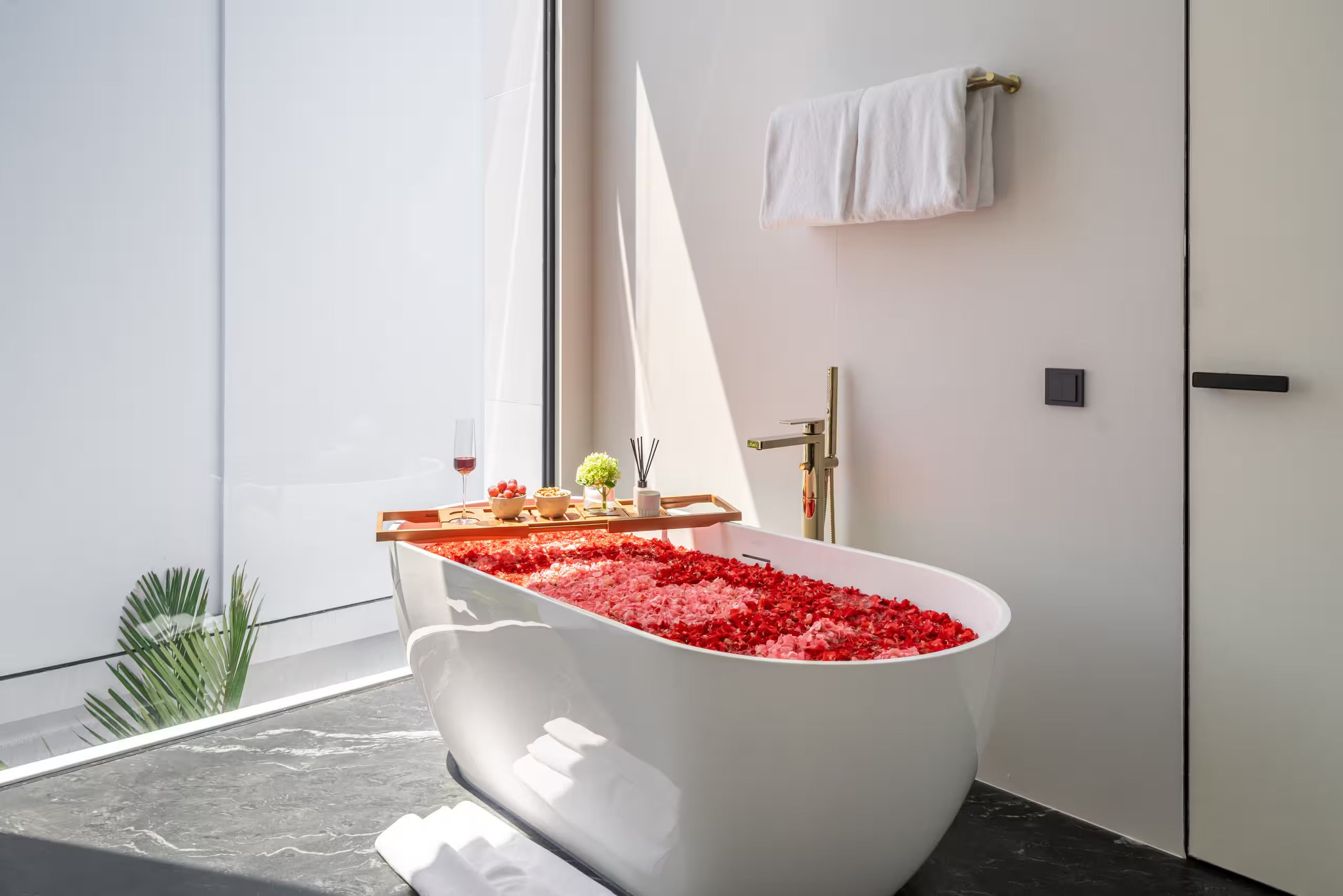 A modern bathroom with a freestanding bathtub filled with rose petals, a towel rack, and a small table with a wine glass and flowers.